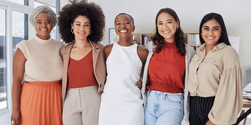 The image shows a group of women smiling. They are standing indoors and wearing casual clothing such as tops, trousers, and jeans. The women are posing for the photo and appear to be happy.