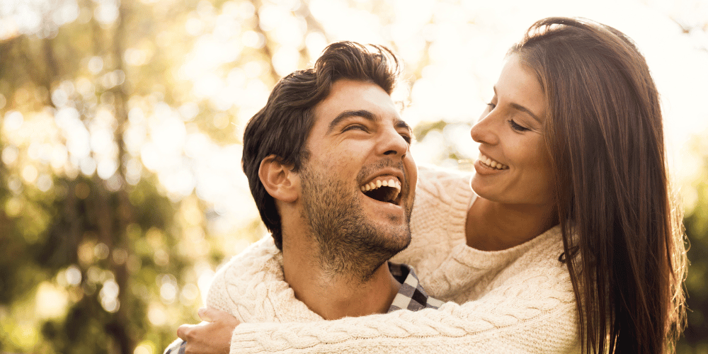 The image features a man and a woman laughing together outdoors. They appear to be enjoying each other's company in a romantic setting.