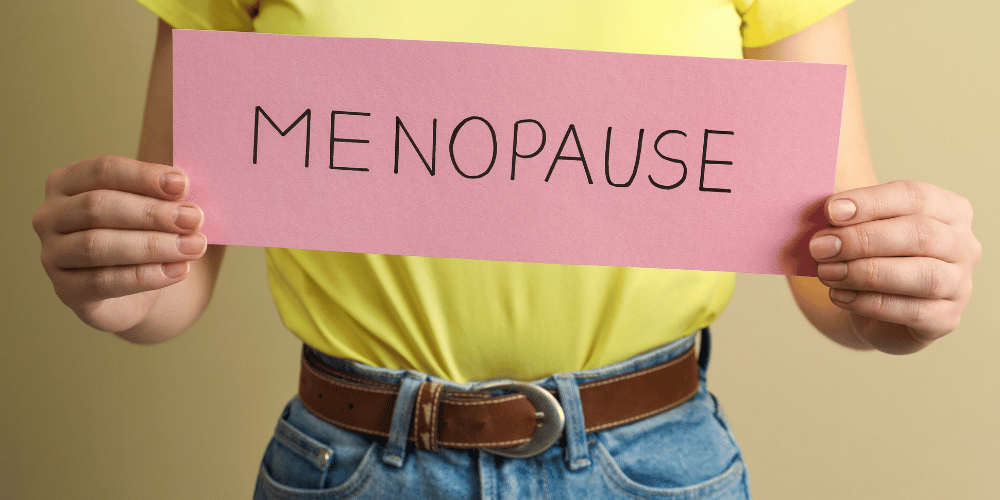 woman with yellow shirt tucked into blue jeans with brown belt holding cardboard pink menopause sign 