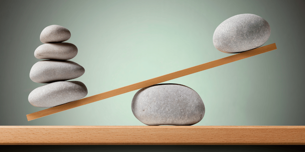 The image shows a wooden shelf with two wooden sticks, one of which has a rock on top. It appears to be a piece of art displayed indoors on a wall.