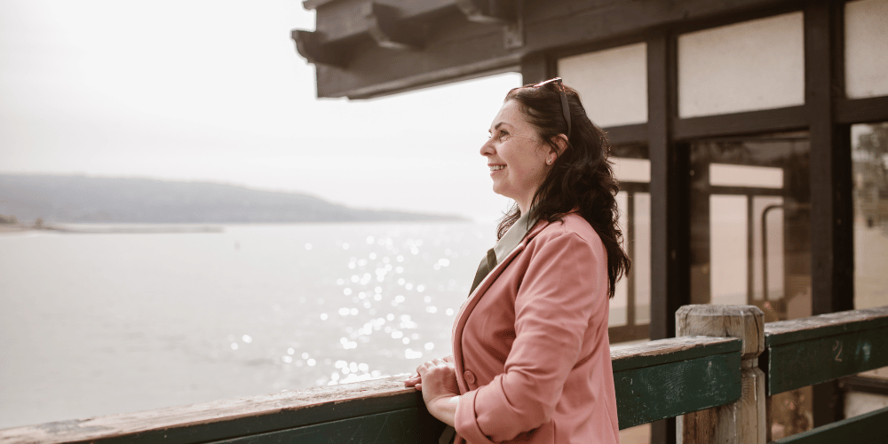 The image shows a person leaning on a railing near a lake. The person is wearing clothing and the background includes water, the sky, and a wooden railing.