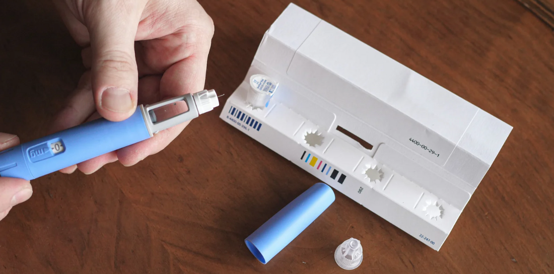 A hand holding a blue injection pen near a white packaging with a needle cap and a blue cap.