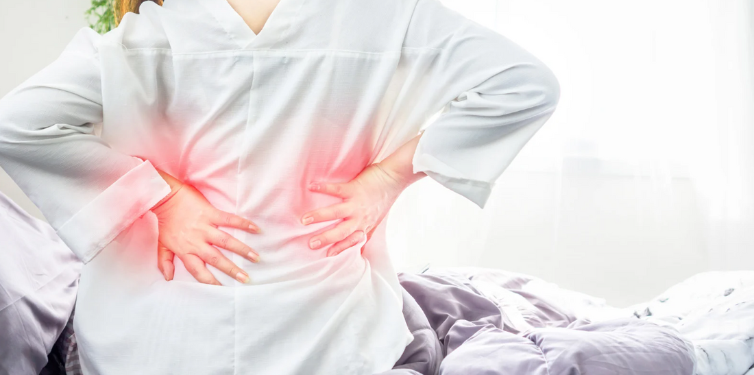 A person is sitting on an unmade bed, wearing a white button-down shirt, with their hands on their lower back.
