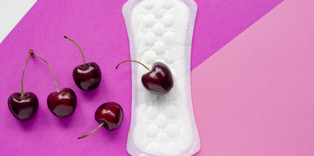 Several dark red fresh cherries are shown on an angled, magenta and pink surface, along with a sanitary napkin.