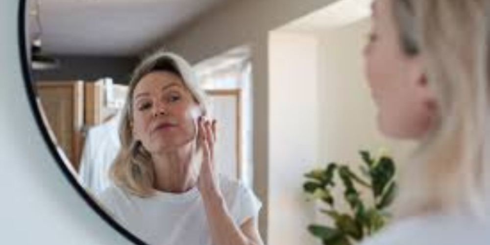 The image shows a woman looking at a mirror indoors. She is likely wearing clothing and standing against a wall.