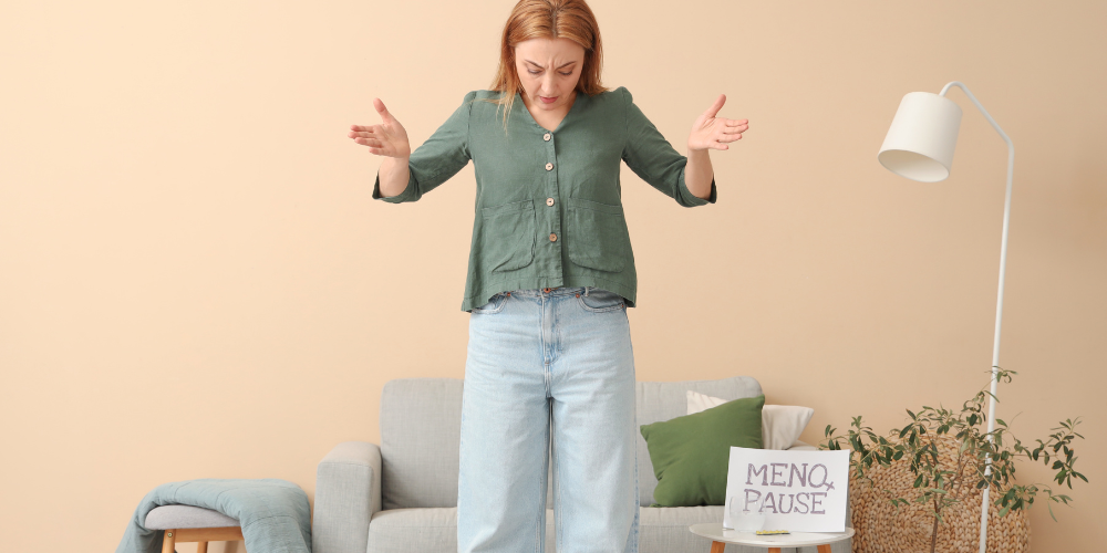 The image features a person standing in a room. The person is wearing jeans and standing next to a couch with a remote controller on it. There is a houseplant in the background as well.