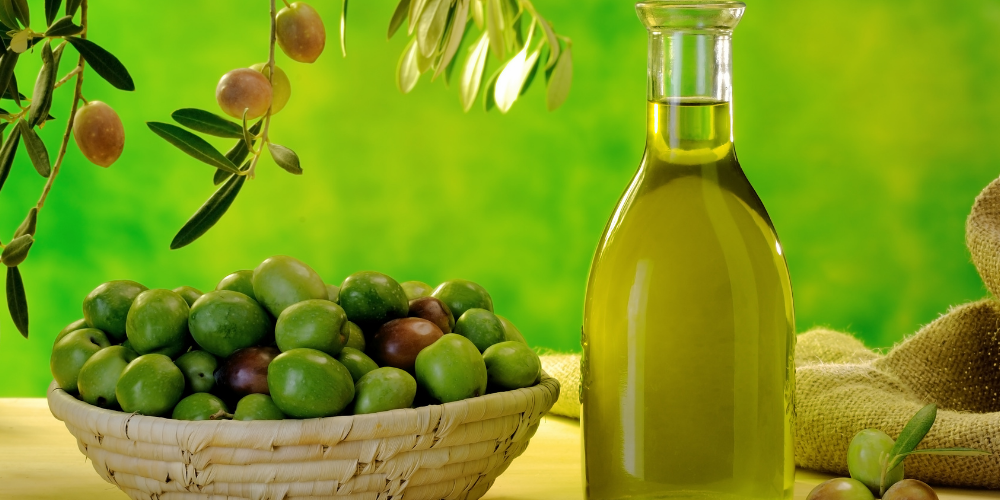 The image is a still life photograph of a basket filled with green olives. The background includes a bottle and a glass of oil..