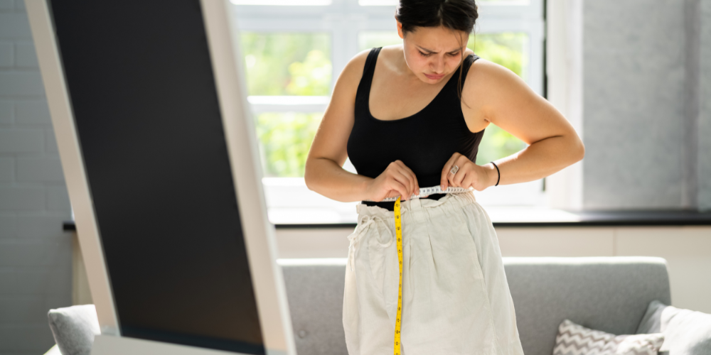 The image shows a woman standing indoors wearing a tank top. She is standing in front of a wall and there is a window visible in the background. The woman's shoulder and elbow are also part of the image.