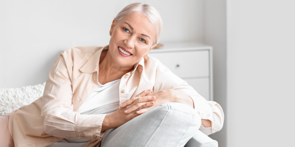 The image shows a woman sitting on a chair indoors. She is smiling and appears to be in a comfortable setting.