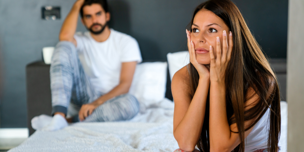 The image shows a man and a woman sitting on a bed with their hands on their faces. The woman has long hair and the setting appears to be indoors, possibly in a bedroom.