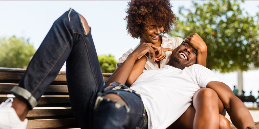 The image depicts a man and woman laughing in a park setting. The man and woman are sitting on a bench under a tree, enjoying a lighthearted moment together.