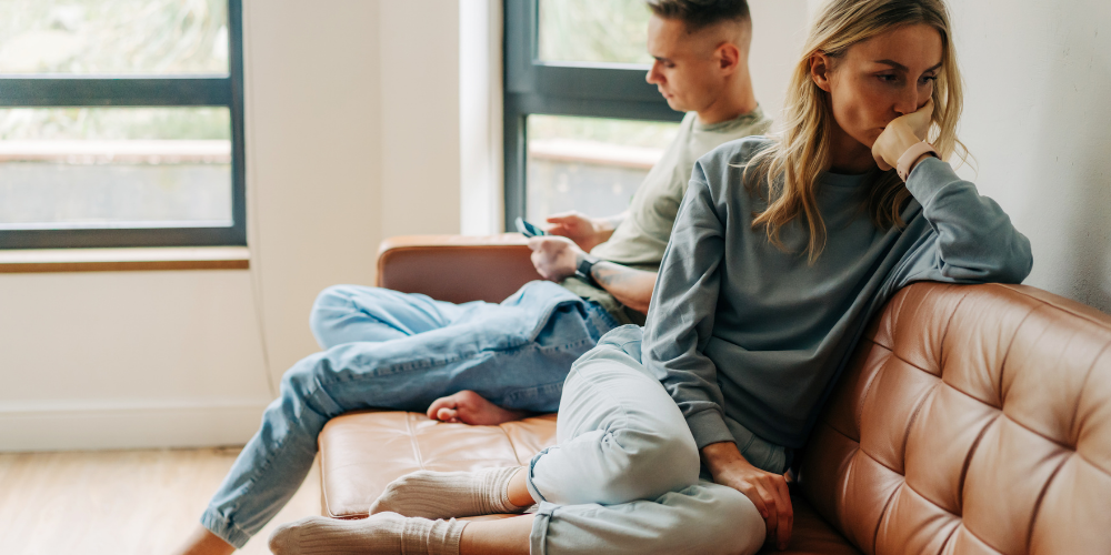 The image shows a man and a woman sitting on a couch indoors. They seem to be tensed around each other in a homely setting. The background includes a wall and a window.