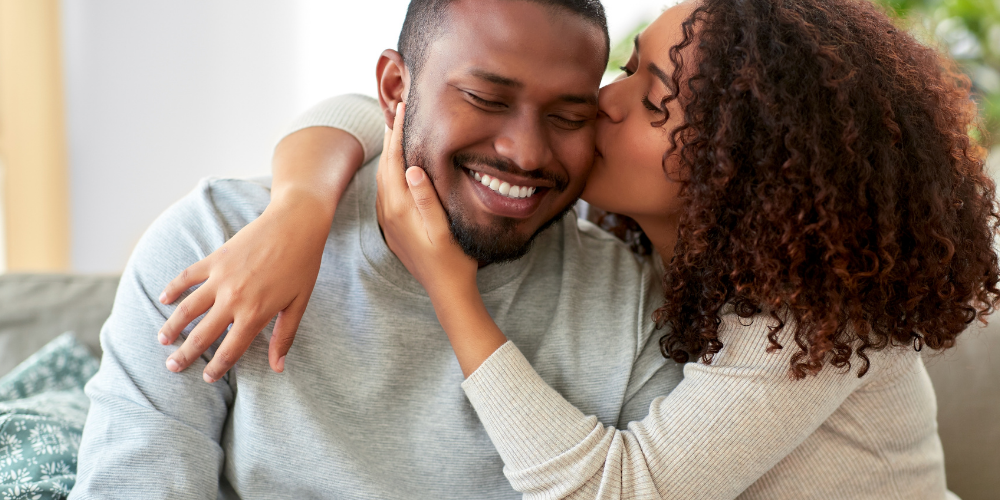 The image depicts a man and woman kissing. The couple is showing affection and love towards each other. They appear happy and are indoors against a wall.