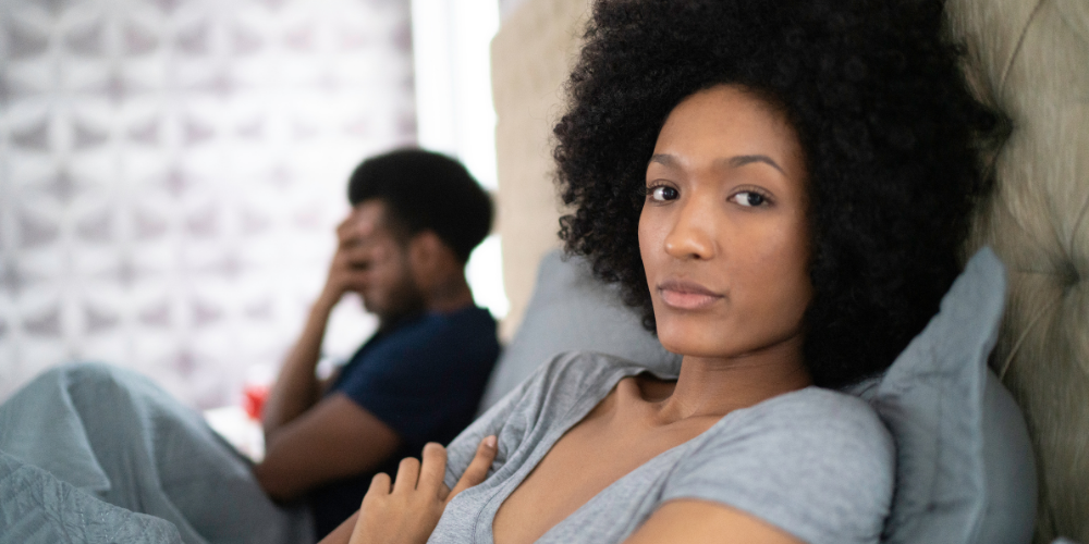 The image features a woman with black hair and a Jheri curl hairstyle looking directly at the camera. She is indoors, standing in front of a wall. The woman appears to be an adult and is dressed comfortably.