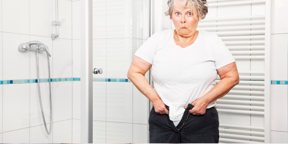 The image shows an old woman squeezing into her jeans inside the bathroom.