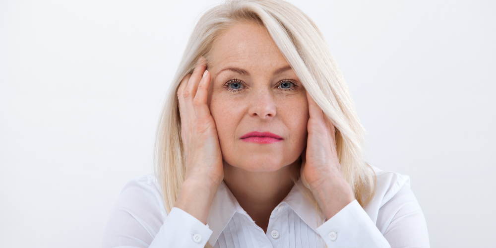 The photo is of a woman with her hand on her face. It is a portrait photograph showing the woman's facial features and clothing. The woman has layered blond hair and is standing against a wall.