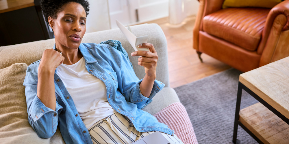 The image is of a woman sitting on a couch indoors. She is smiling and appears comfortable. The background shows a wall and a pillow on the couch.