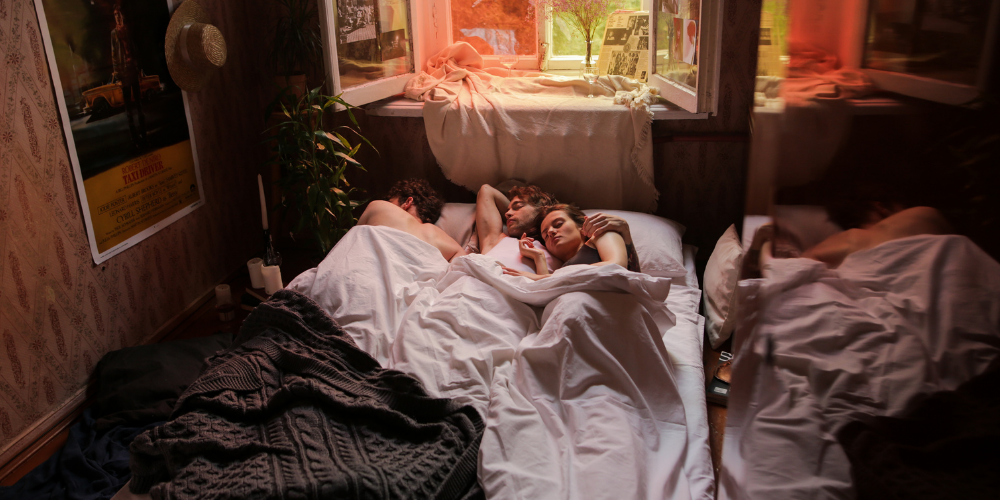 The image shows a couple of people lying in a bed. The scene is indoors, featuring a bed, human faces, a wall, a window, and a room conveying comfort.