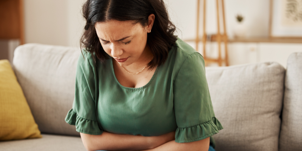 The image shows a woman sitting on a green couch indoors. She is wearing a green shirt and appears to be comfortable with a pillow beside her.