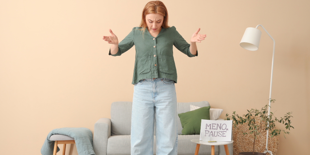 The image shows a person standing in a room. The person is wearing jeans and is standing next to a couch and a table. There is a houseplant in the room as well.