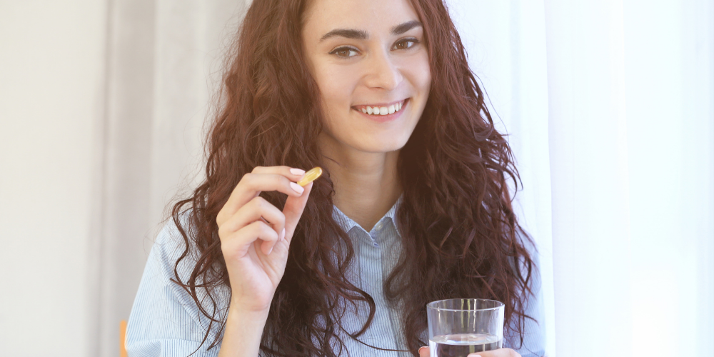 The image shows a woman holding a pill and a glass of water indoors.