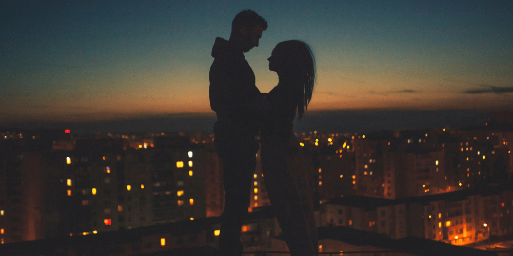 The image shows a silhouette of a man and woman kissing in front of a city at night.