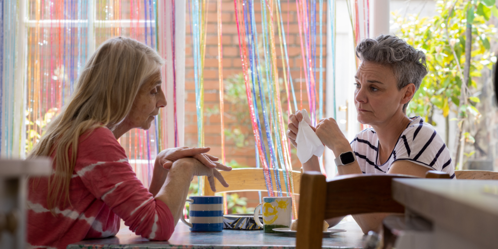 The image shows two women sitting indoors and having a conversation.