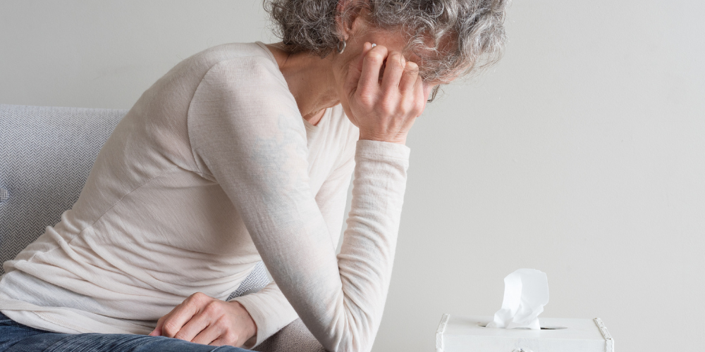 A woman is sitting indoors, holding her face with her hands. She appears to be in a moment of reflection or contemplation. The setting includes a wall in the background, and she is dressed in comfortable clothing.